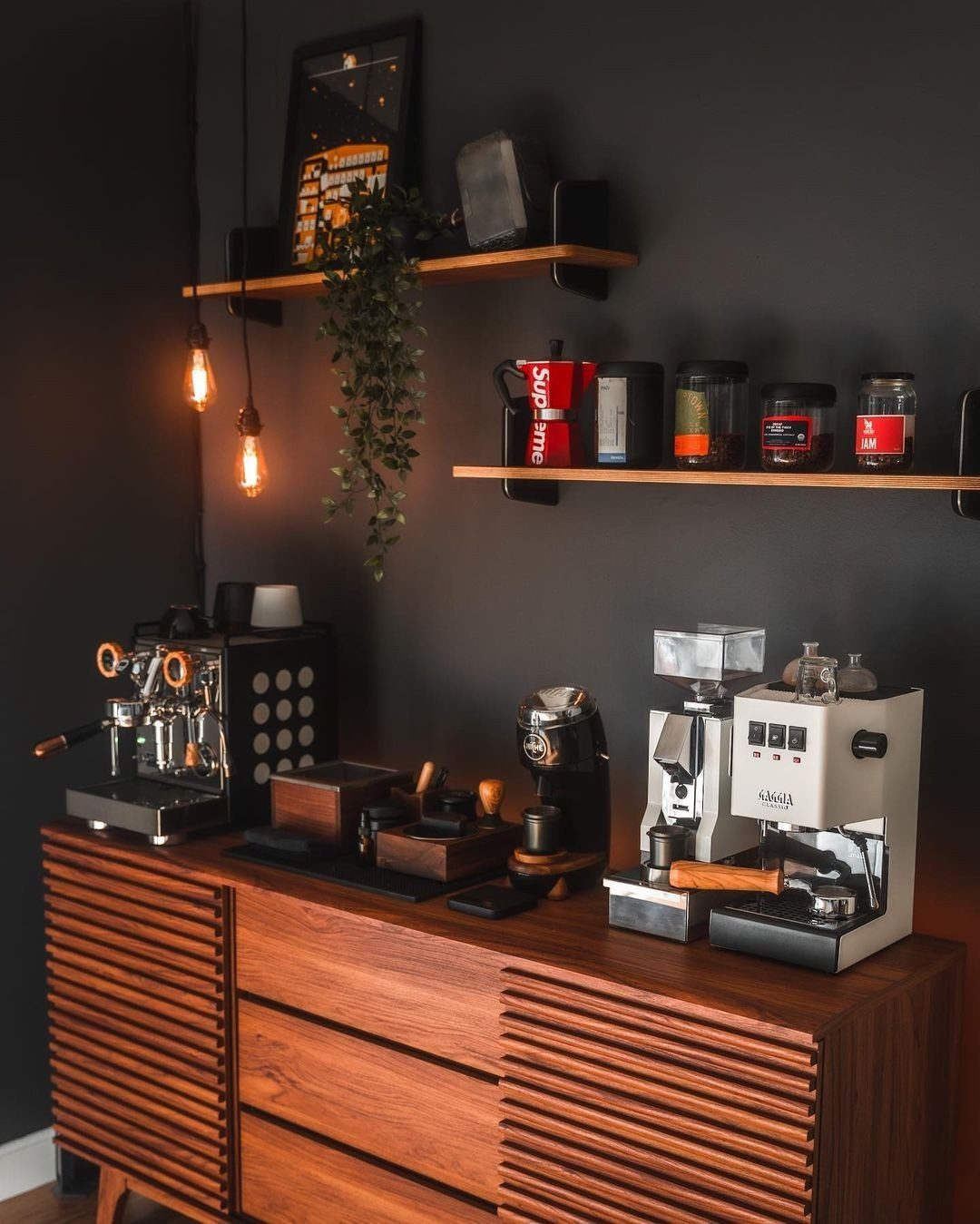 Two espresso machines and coffee accessories arranged on a wooden sideboard under floating shelves.