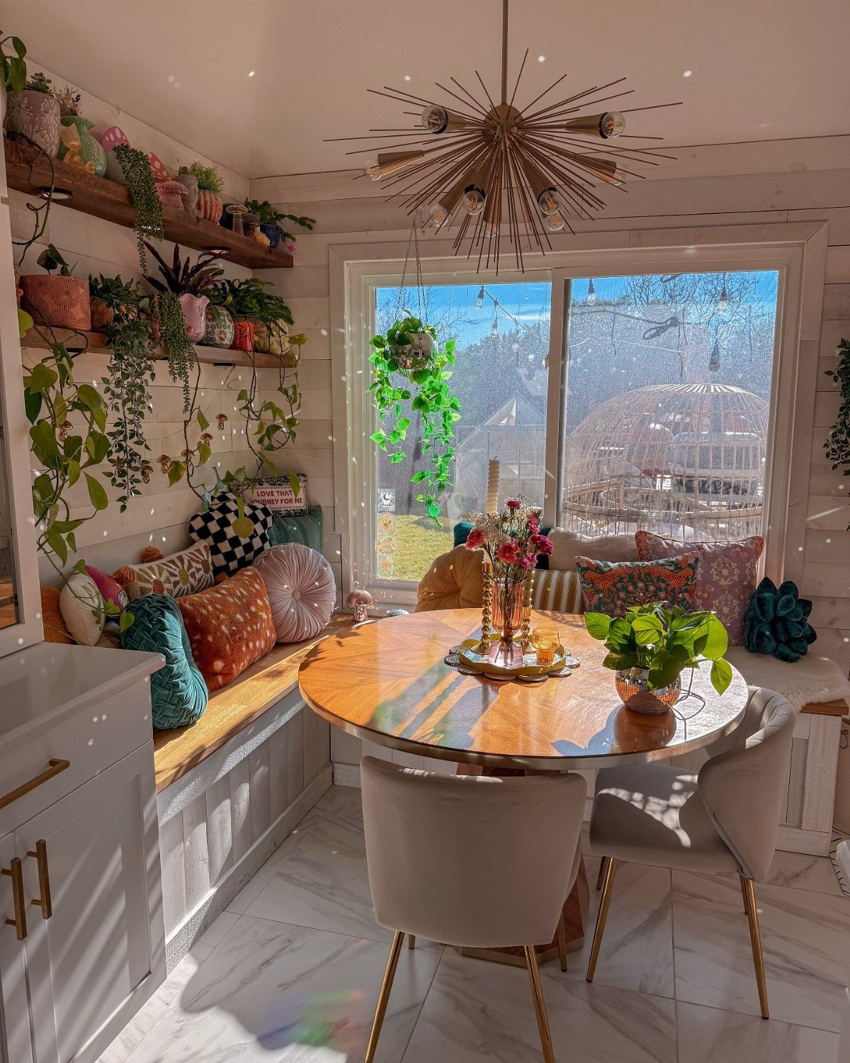 20 Simple and Small Kitchen Design Ideas 20 Round wooden dining table with chairs and a cushioned window bench surrounded by green plants.