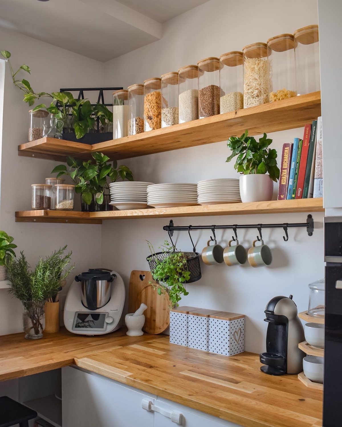 20 Sleek Kitchen Cabinet Design Ideas for 2026 12 Wooden open shelves holding glass jars, white plates, plants, and books against a white wall.
