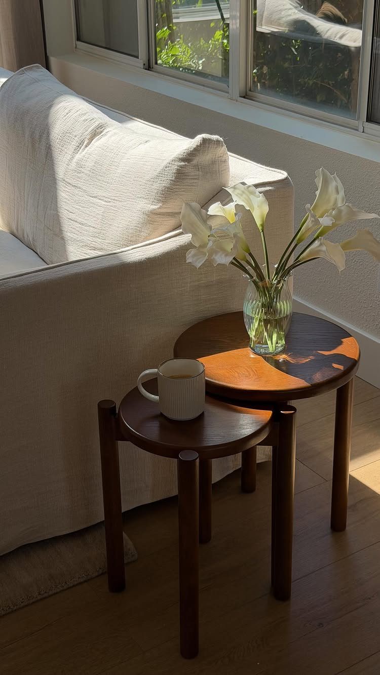 Two round wooden nesting tables with flowers and a mug next to a white sofa.