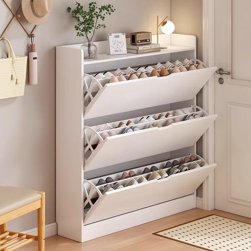 White slim shoe rack with three tilting compartments filled with shoes against a wall.