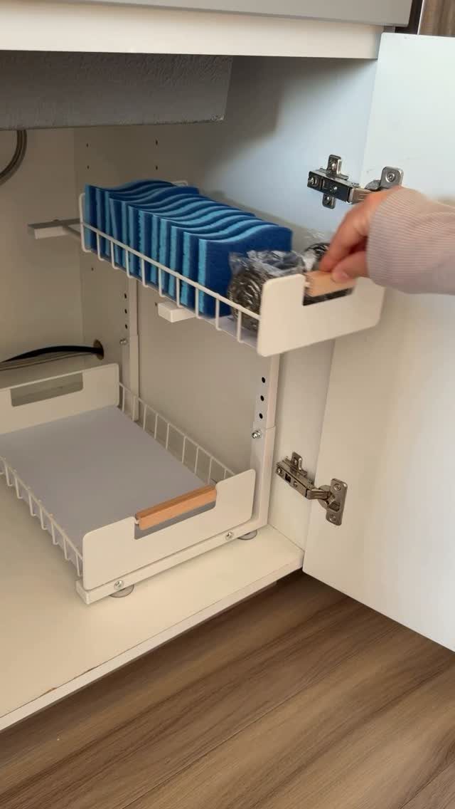 14 Small Apartment Storage Hacks and Genius Finds for 2026 9 A hand pulls out a white sliding drawer organizer containing blue cloths inside a kitchen cabinet.