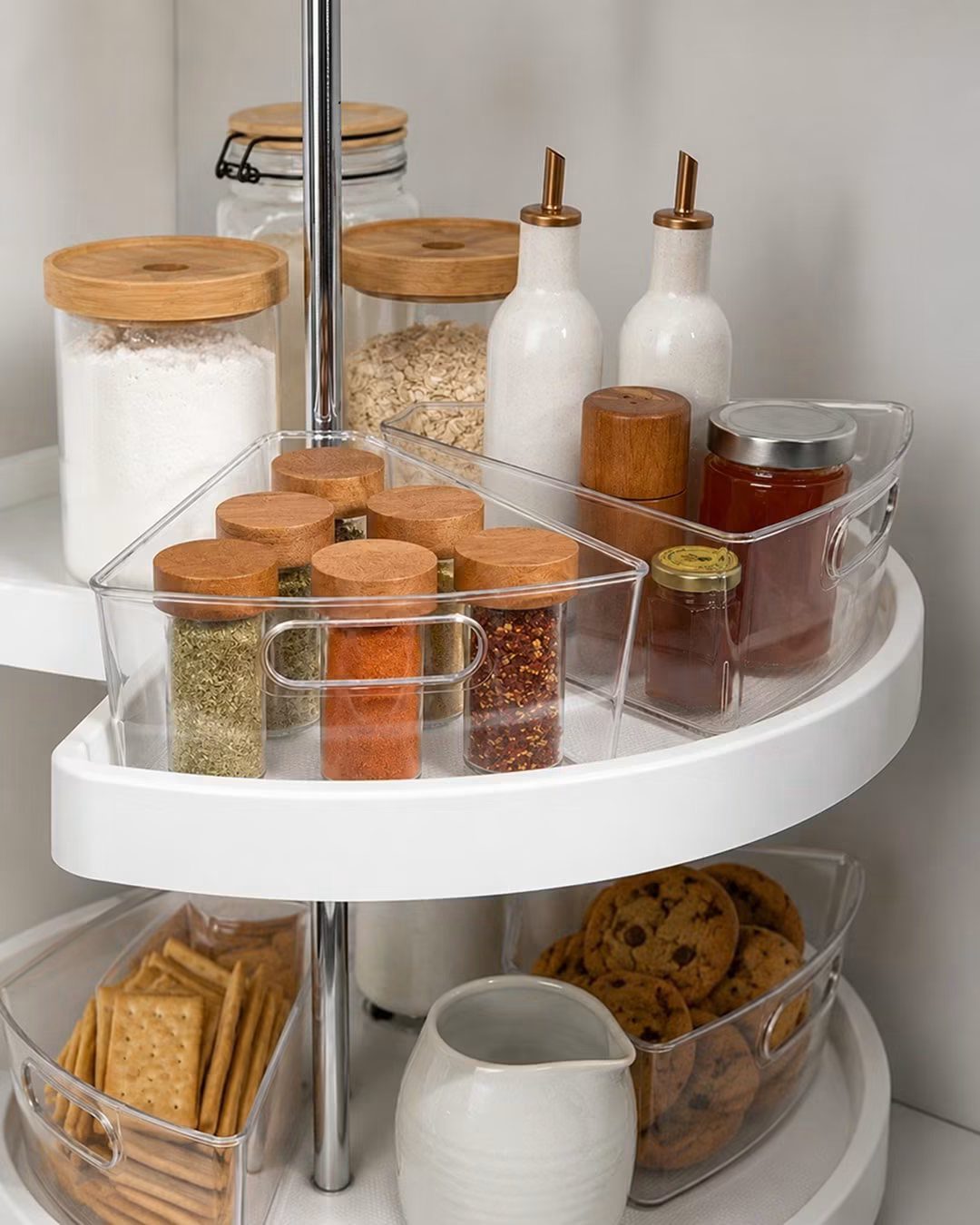 A white two-tier rotating organizer shelf holding various spices, jars, and snacks in a pantry.