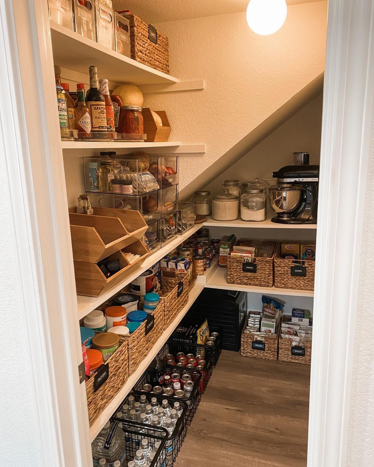 An organized under-stair pantry featuring white shelves, wicker baskets, clear containers, and a stand mixer.
