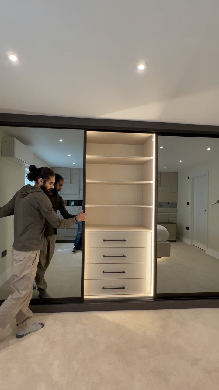 A man opens a large sliding mirrored wardrobe containing white shelves and drawers.