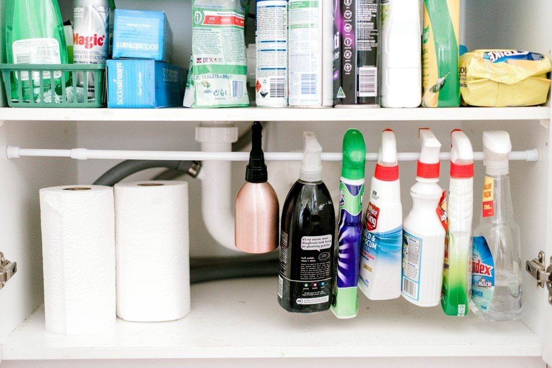 White tension rod holding cleaning spray bottles under a bathroom sink cabinet.