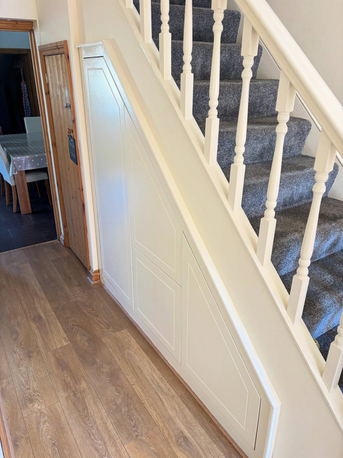 Cream paneled bespoke cabinets installed underneath a carpeted staircase in a home hallway.