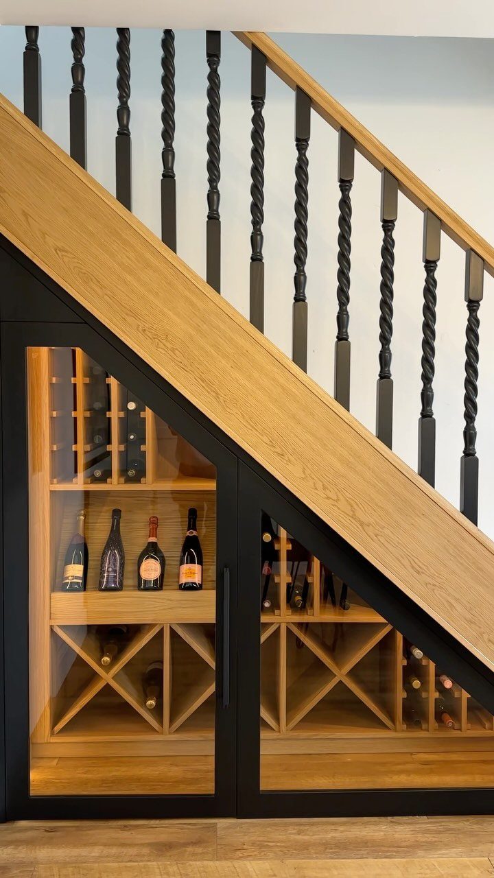 Wooden wine rack with glass doors installed under a staircase with black spindles.